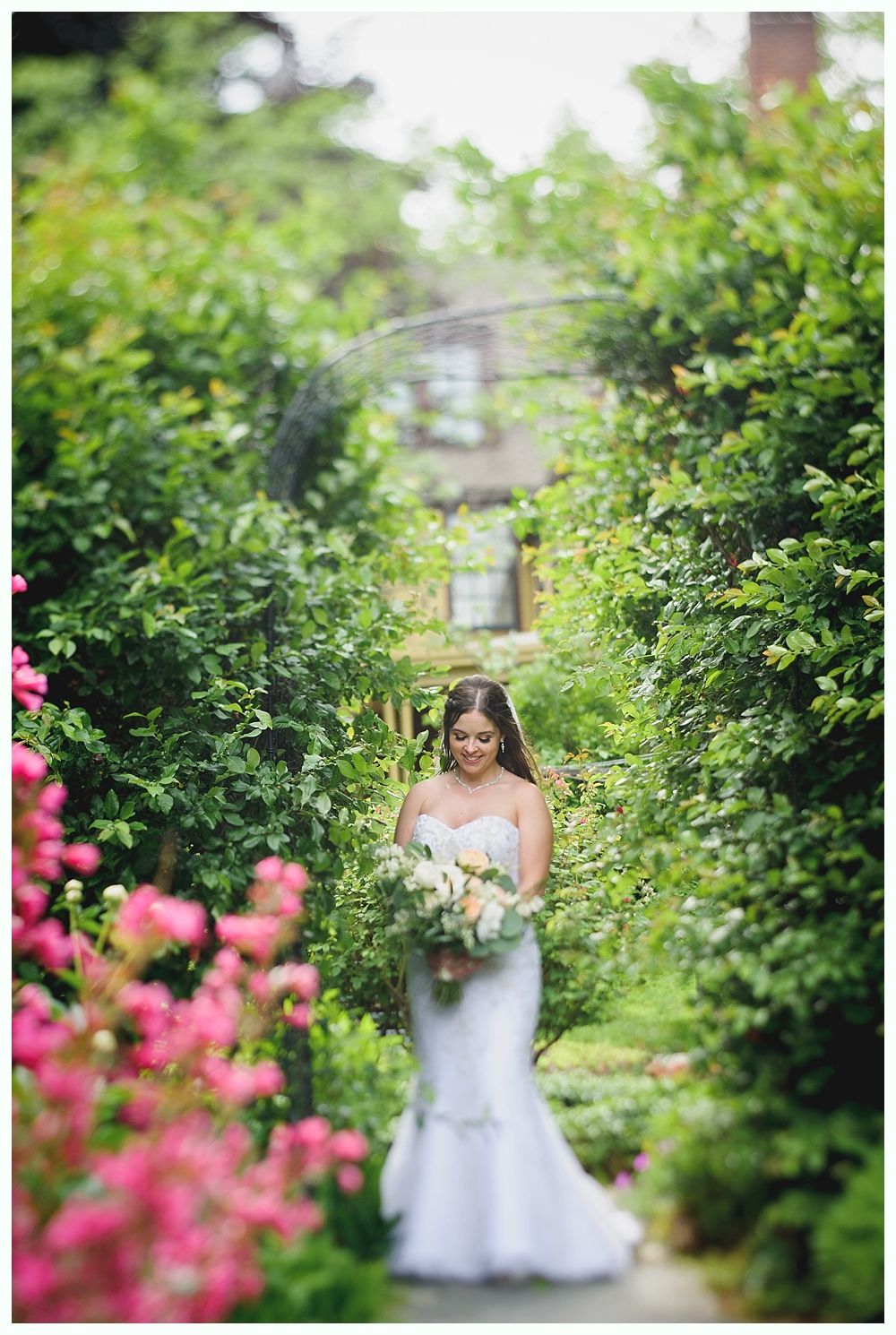 Bride in white gown holding flowers, standing in a lush, green garden pathway.
