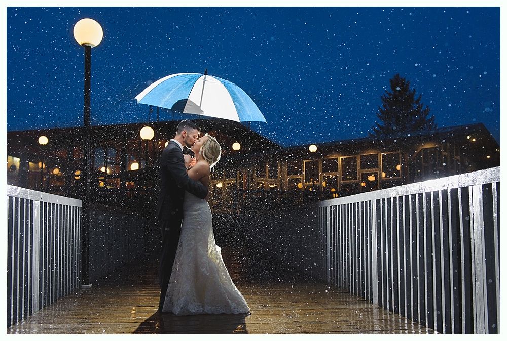 Couple kissing under umbrella on a bridge at night; rain falling, lights glowing.