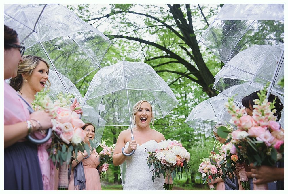 Bride and bridesmaids in dresses, smiling, holding bouquets and umbrellas, outdoor setting.