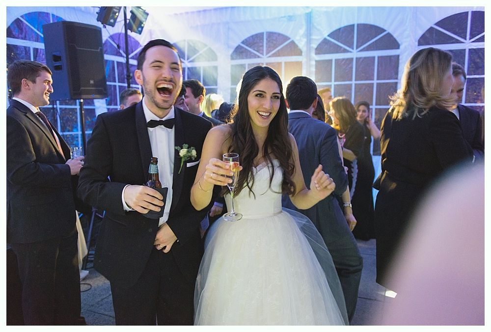 Bride and groom dancing at a wedding reception, laughing and holding drinks. Festive tent in the background.