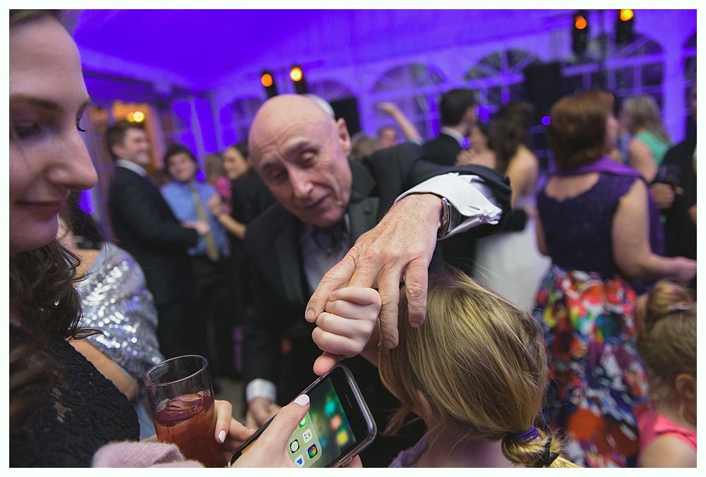 Man in tuxedo adjusts a girl's hair while someone takes a photo with a phone at a party.