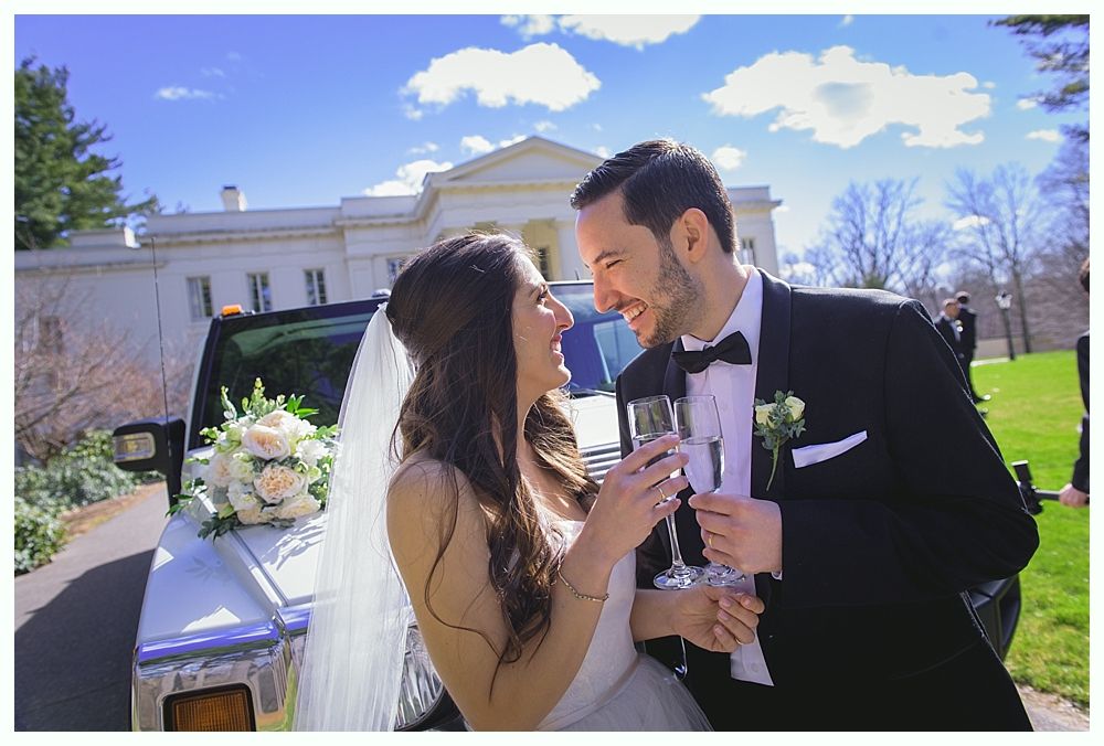 Bride and groom toast with champagne in front of a white building, sunny day.