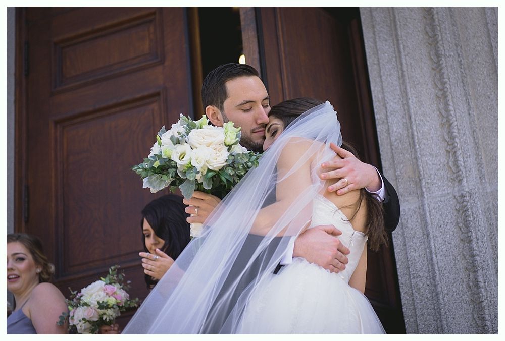 Newlyweds embrace outside a building. Bride in white gown and veil, groom in suit, holding a bouquet.