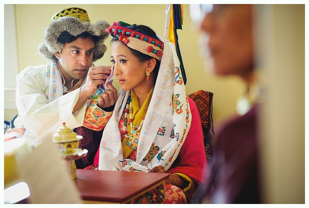 Man adjusts ear of woman in traditional clothing, seated near others, likely at a ceremony.