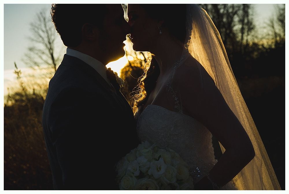 Wedding couple embracing, silhouetted against a setting sun. Bride in veil and dress, holding bouquet.