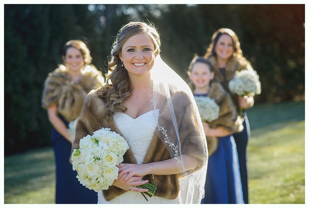 Bride in white dress holding flowers, with bridesmaids in blue dresses and faux fur shawls.