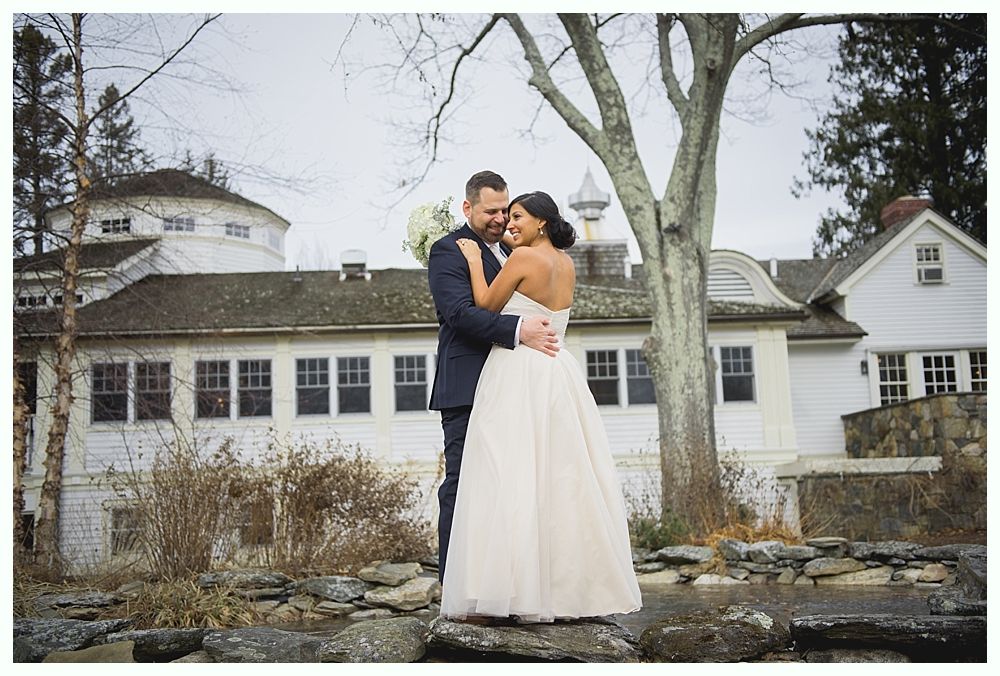Couple embracing in front of a white house with a round roof. The bride wears a strapless gown, the groom a suit.
