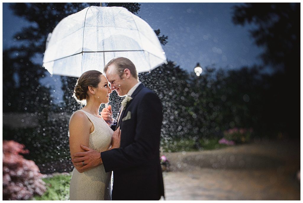 Newlyweds embrace under a clear umbrella in the rain, with a garden setting and soft lighting.