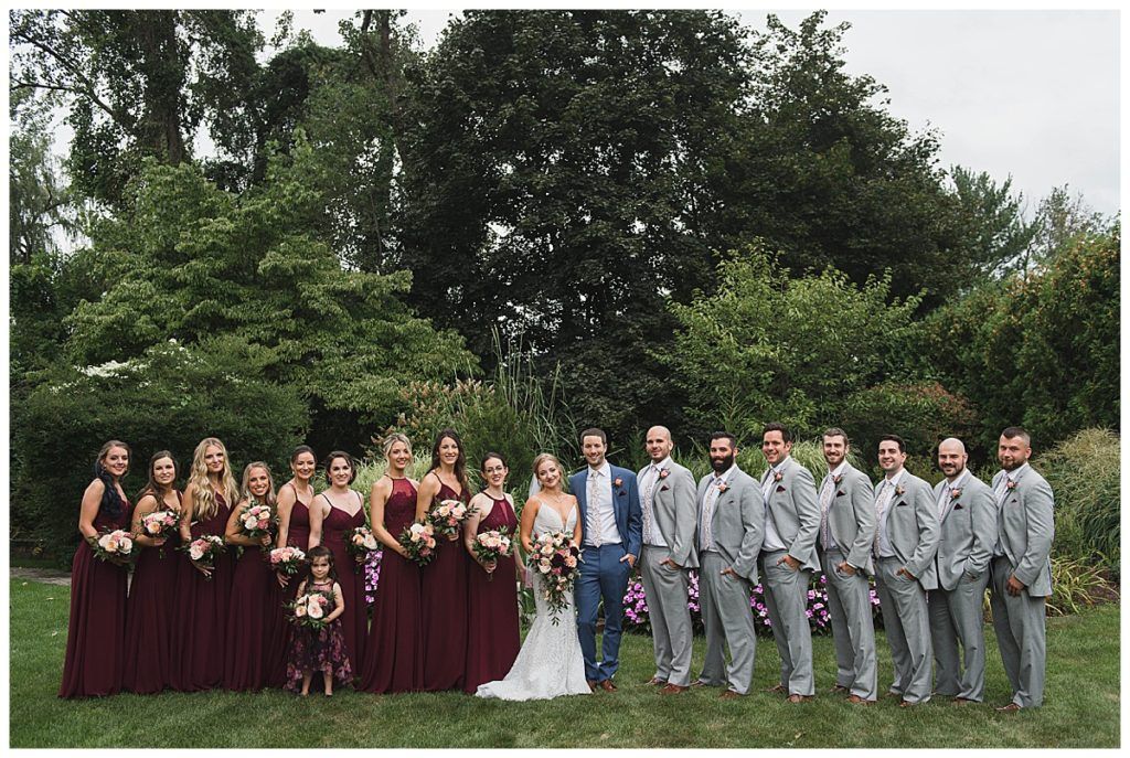 Wedding party poses outdoors, bride and groom in front, bridesmaids in burgundy dresses, groomsmen in gray suits.