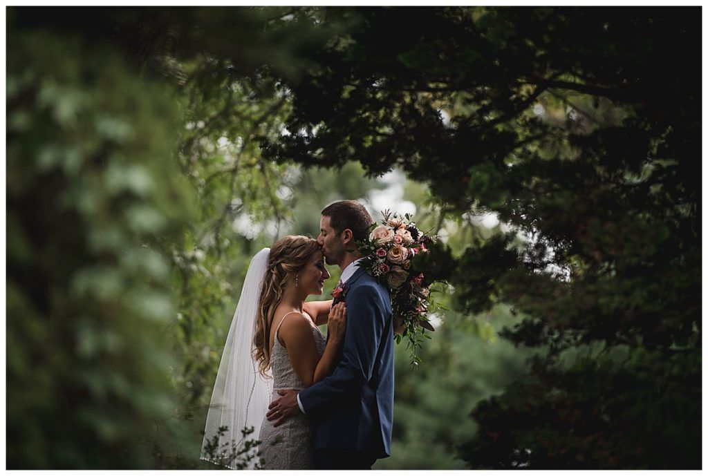 Bride and groom embracing, forehead to forehead, in a forest setting with soft focus.
