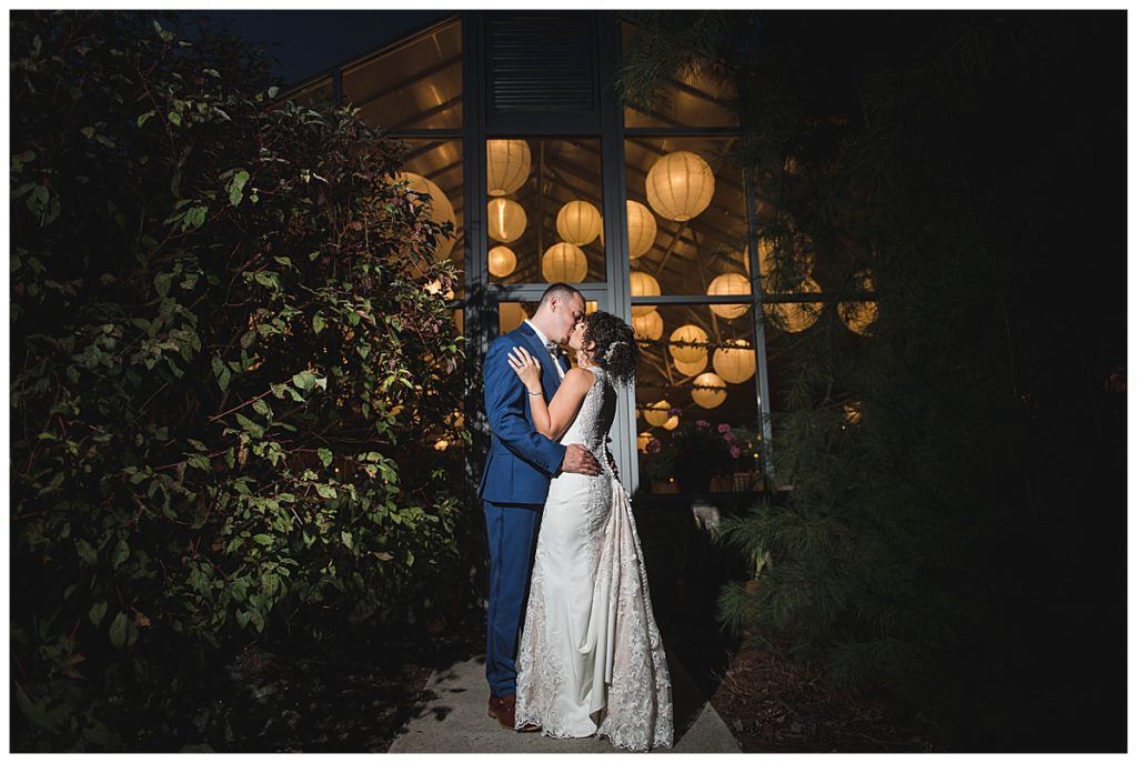 Bride and groom embrace in front of a window lit with paper lanterns at night.