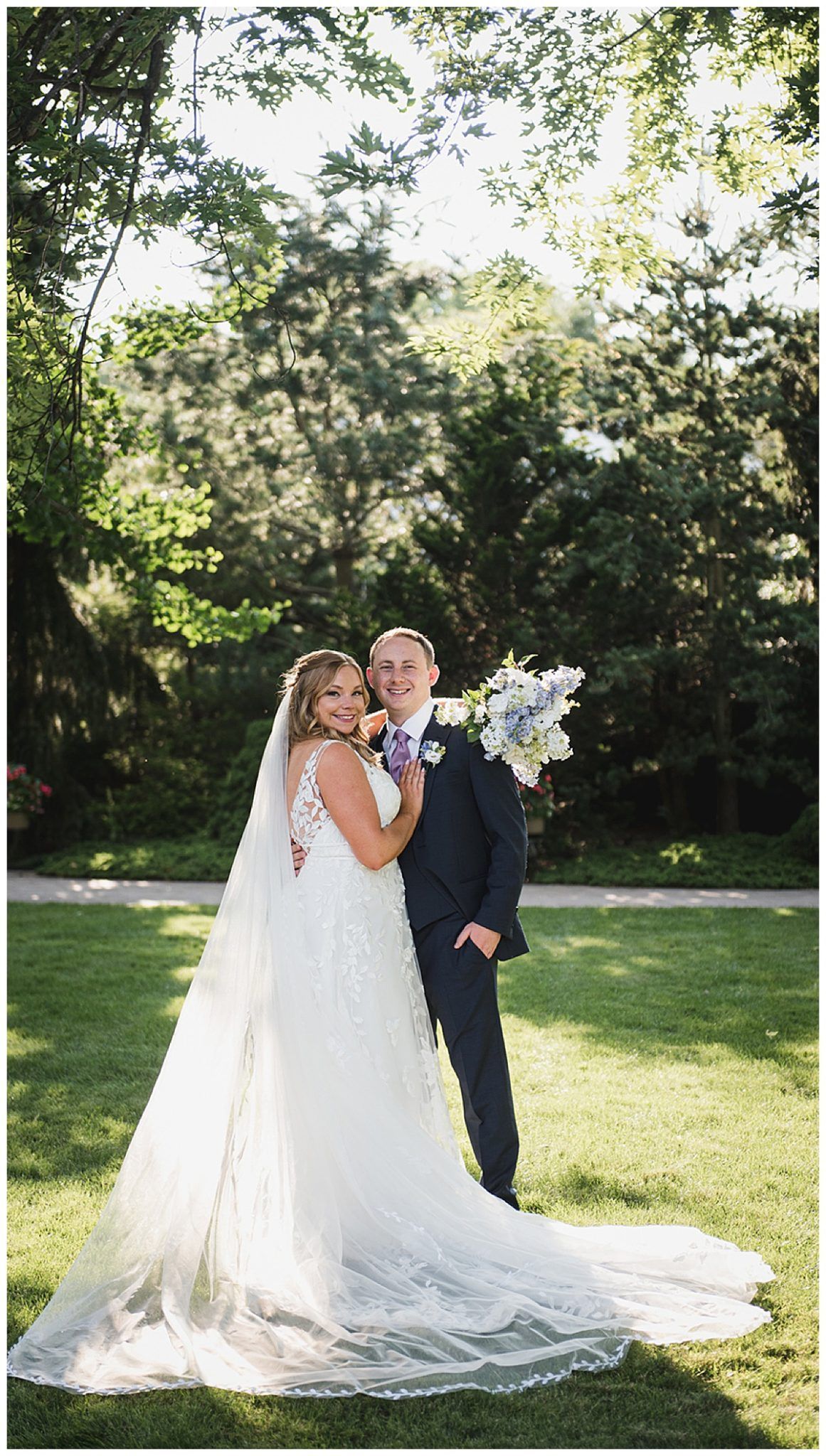 Bride and groom pose on grass. The bride wears a white dress and veil, holding a bouquet. The groom wears a suit. They stand outside near trees.