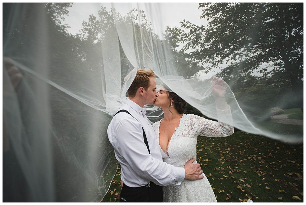 Newlyweds kiss under a flowing white veil in a park setting.
