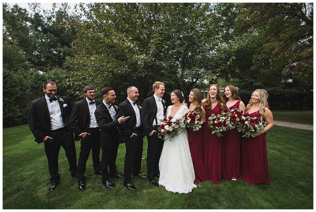 Wedding party poses on grass: bride in white dress, bridesmaids in red, groomsmen in black, joyful expressions.