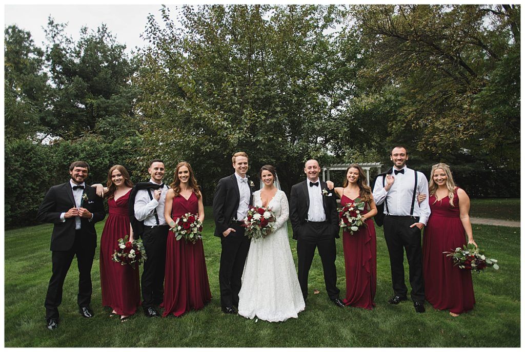 Wedding party poses outdoors. Bride and groom stand with bridesmaids in burgundy dresses and groomsmen in black tuxedos.