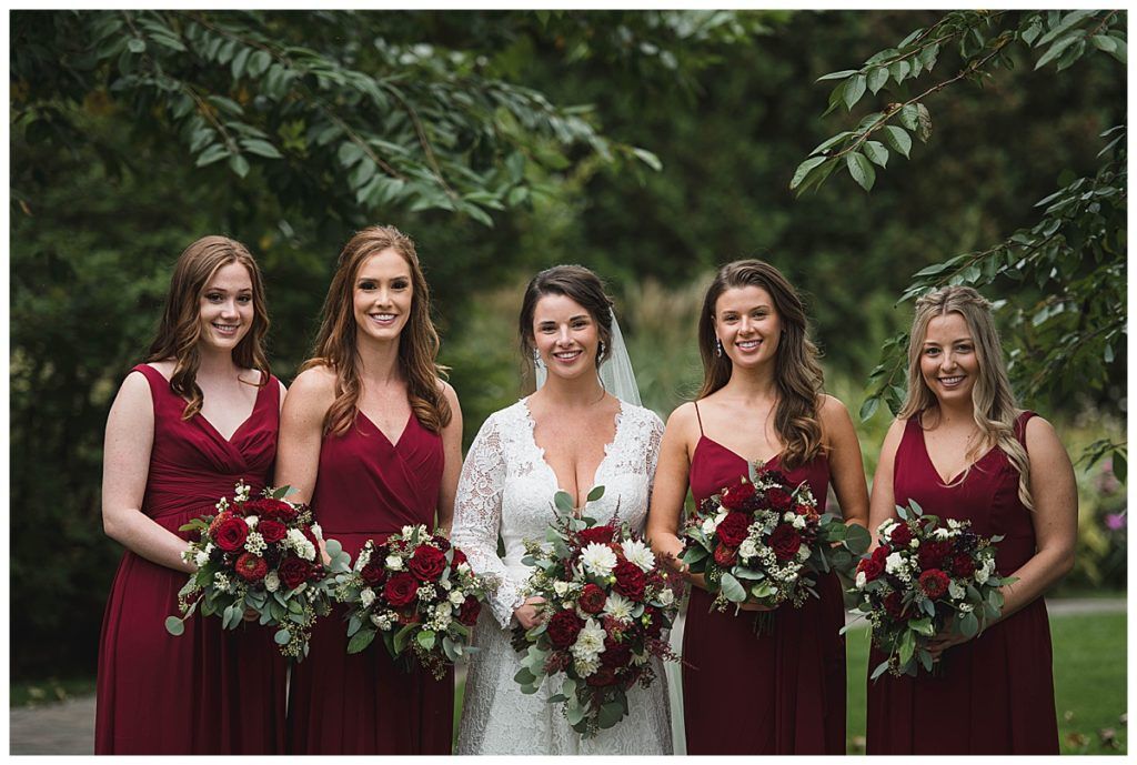 Bride and bridesmaids pose with bouquets; bride in white lace, bridesmaids in burgundy dresses.