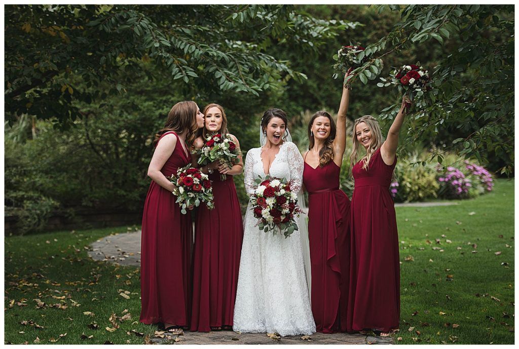Bride with four bridesmaids in burgundy dresses, outdoors, holding bouquets.