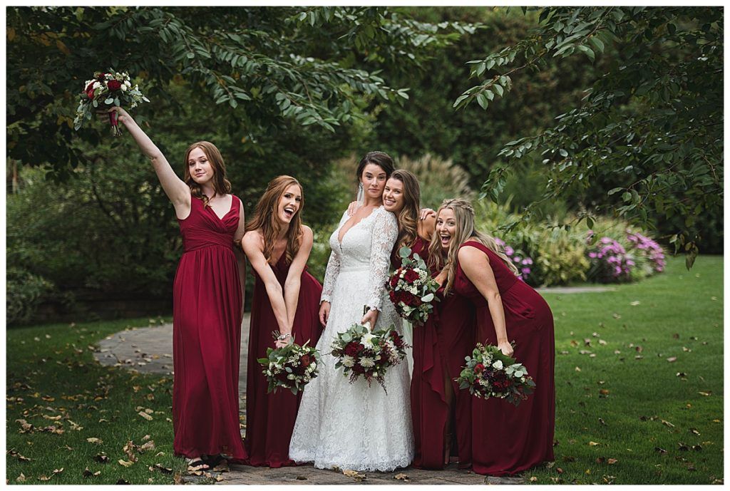 Bride and bridesmaids in burgundy dresses pose outdoors, holding bouquets.