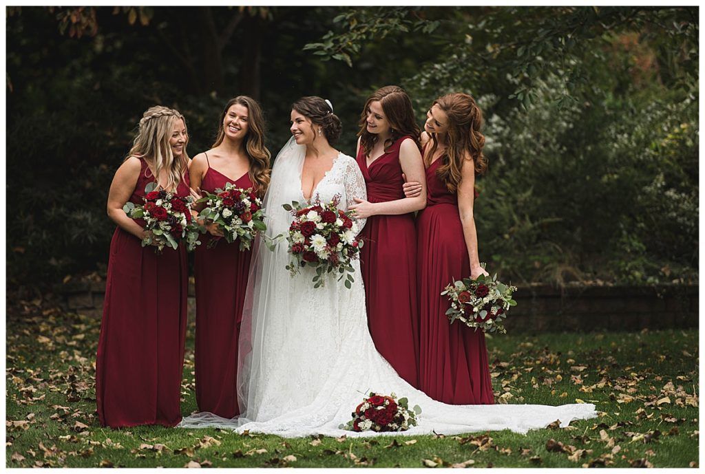 Bride in white dress with bridesmaids in burgundy dresses, holding bouquets outdoors.