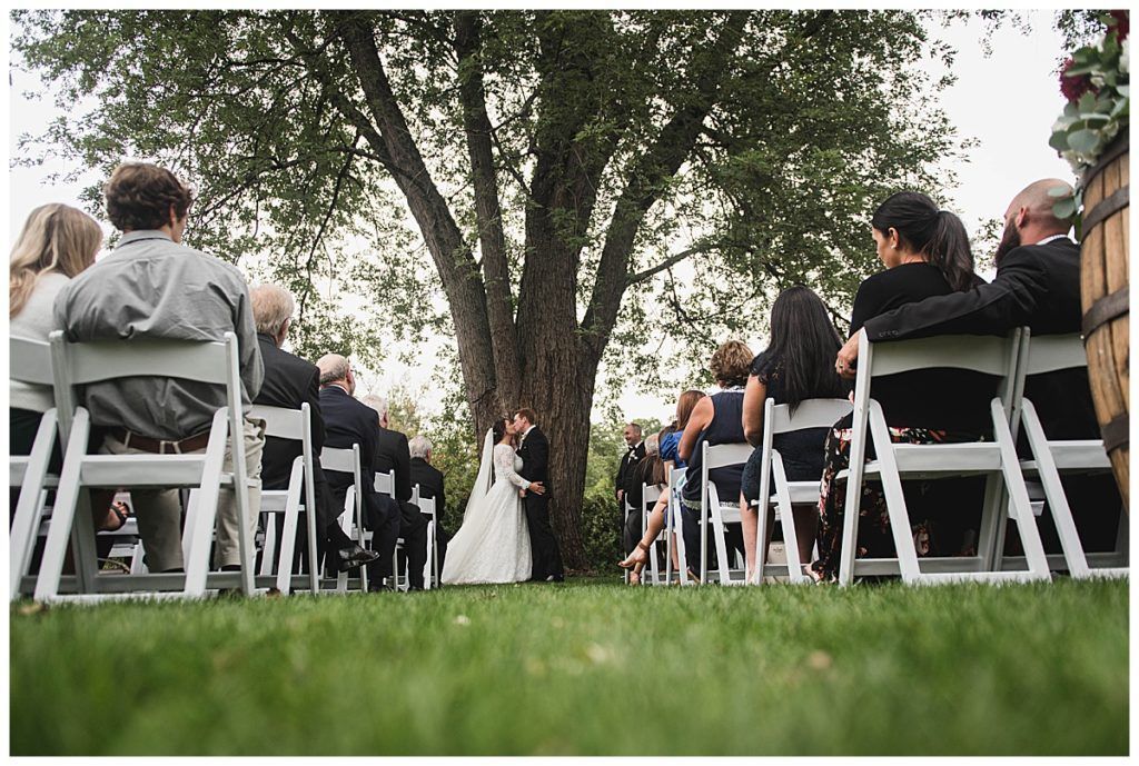 Wedding ceremony: Bride and groom kissing under a tree, guests seated on white chairs.