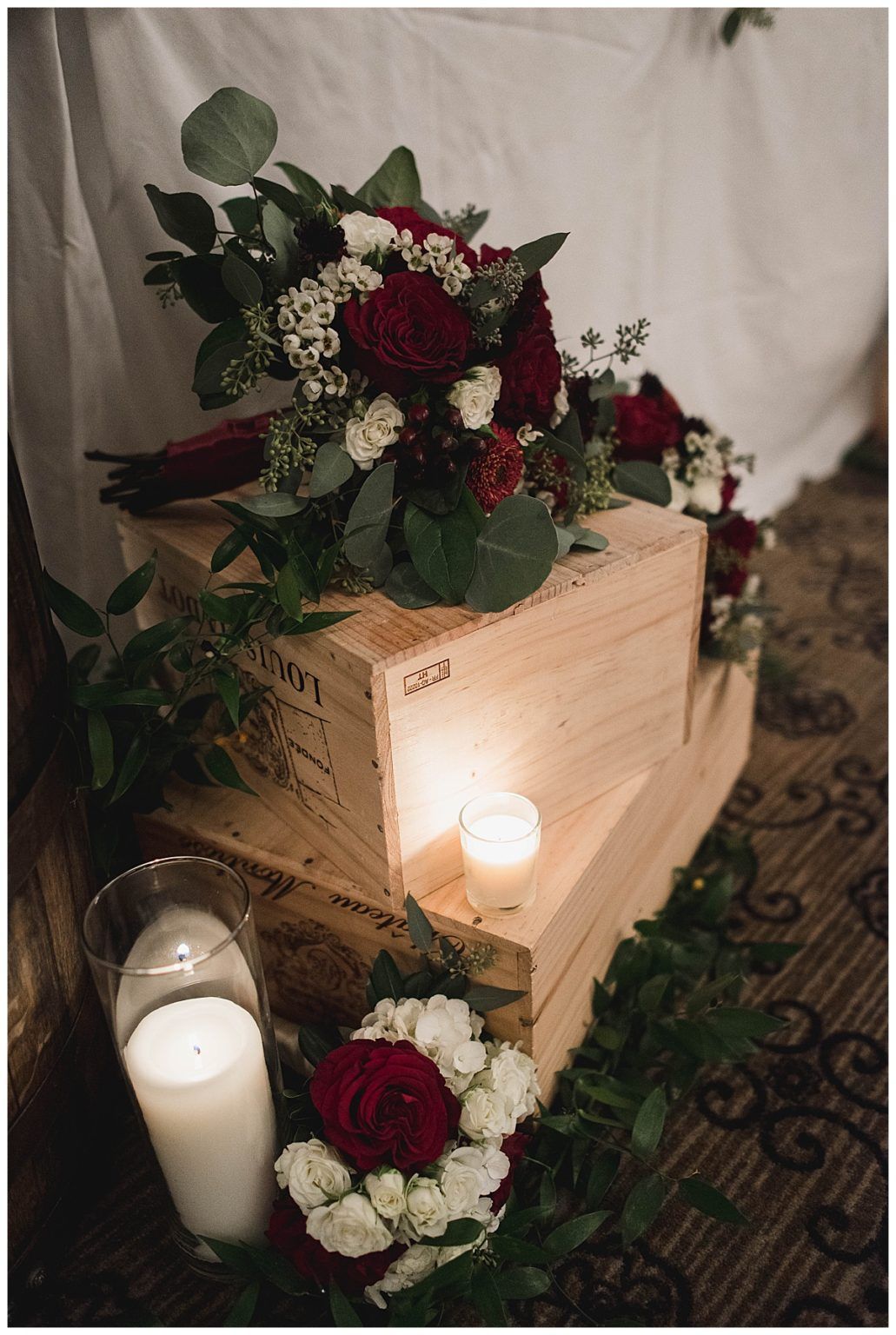 Wedding bouquet with red flowers, greenery, and candles displayed on wooden boxes.