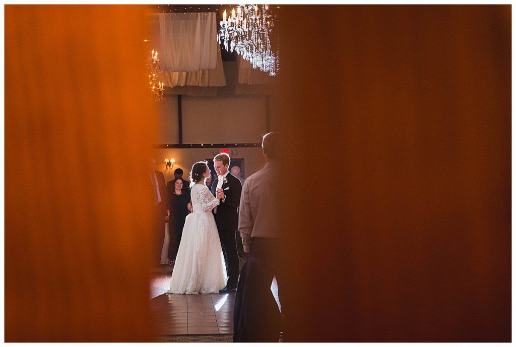 Bride and groom dance at a reception, seen through orange curtains. Guests and chandelier in the background.
