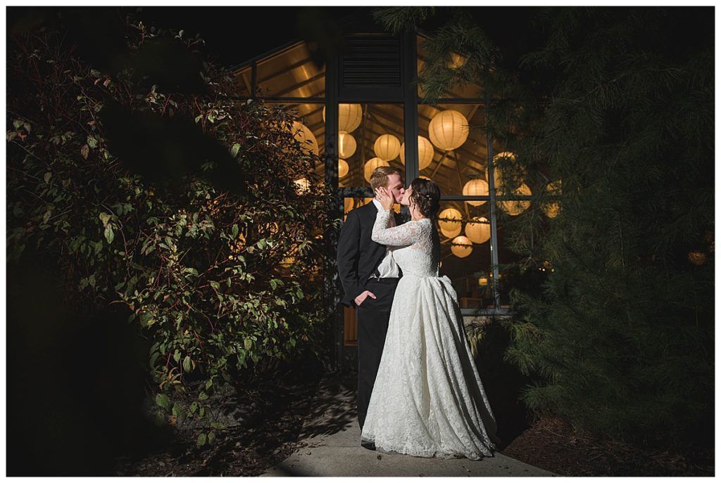 Couple kissing outside a building at night. Bride in a white dress, groom in a tuxedo, soft lights in the background.