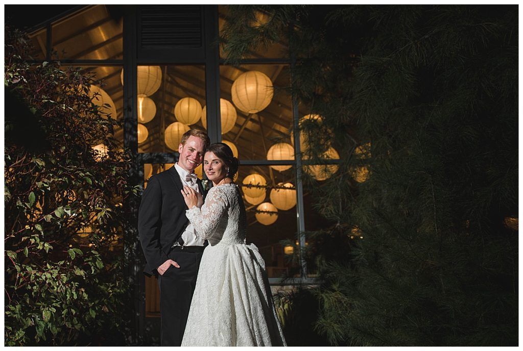 Couple posing by lit lanterns in front of a glass wall. Man in suit, woman in white dress smiling.