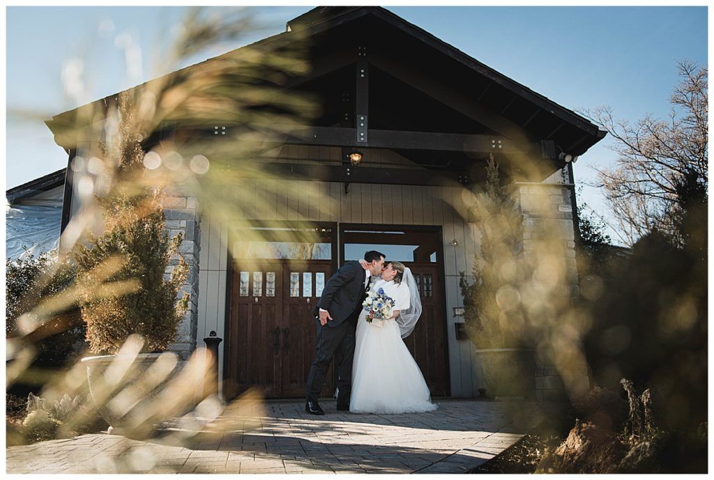 Bride and groom kissing in front of a wooden building with arched entrance.