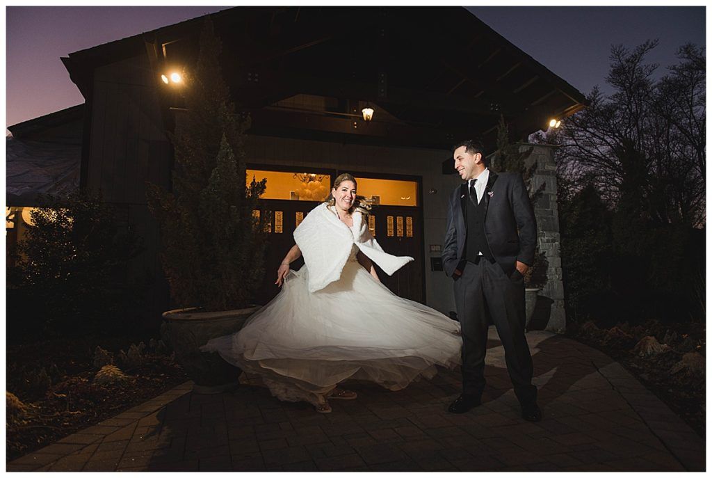 Bride twirls in a white gown and shawl, groom watches on a stone path at night, building in background.