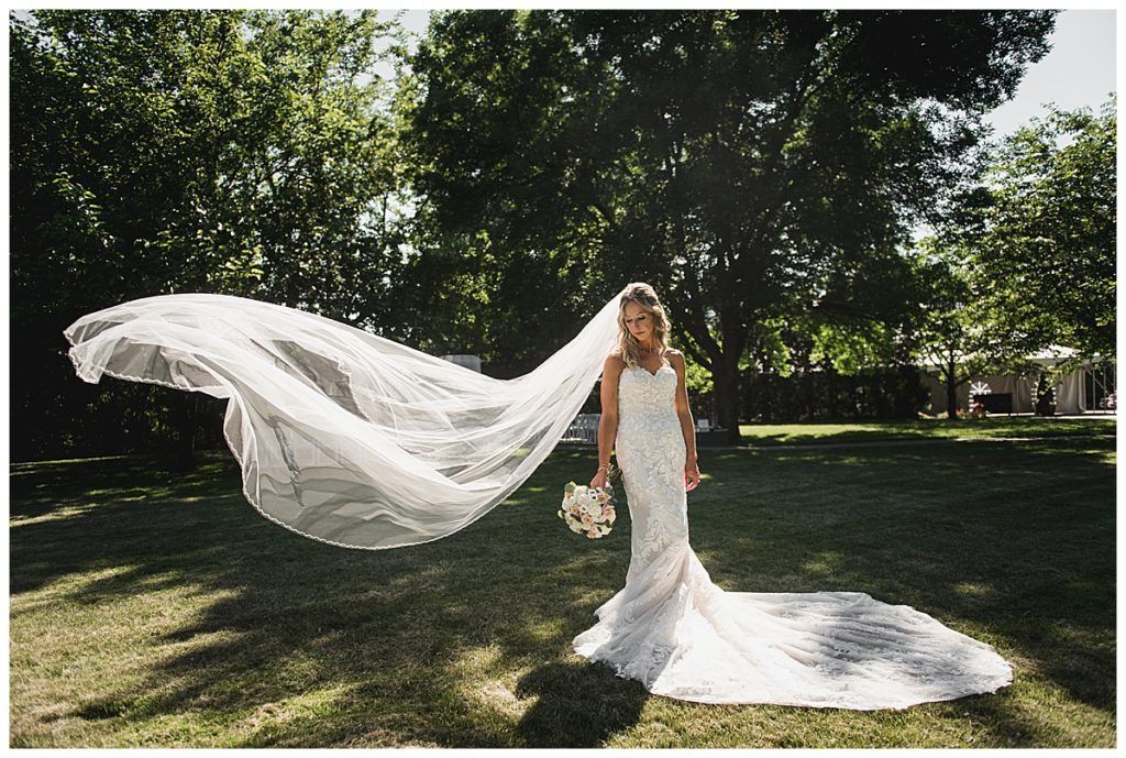 Bride in white lace wedding dress, veil billowing in the wind, holding bouquet, standing on grass.