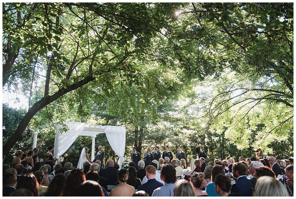 Wedding ceremony under trees; white canopy; guests seated, watching event.