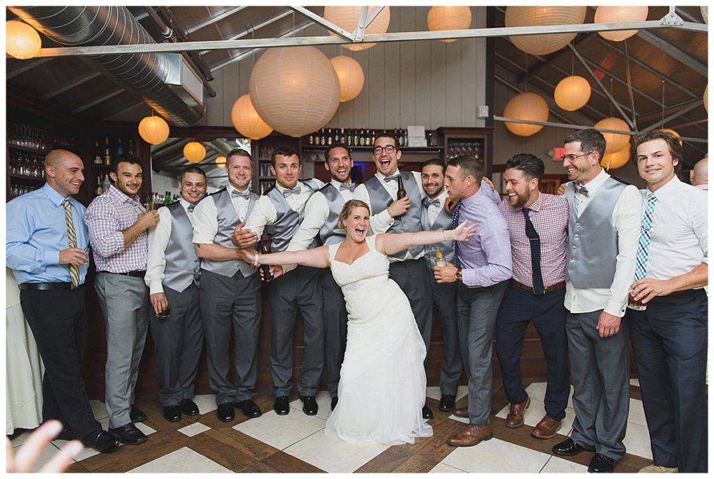 Wedding party posing, bride with arms outstretched, surrounded by groomsmen, festive indoor setting.