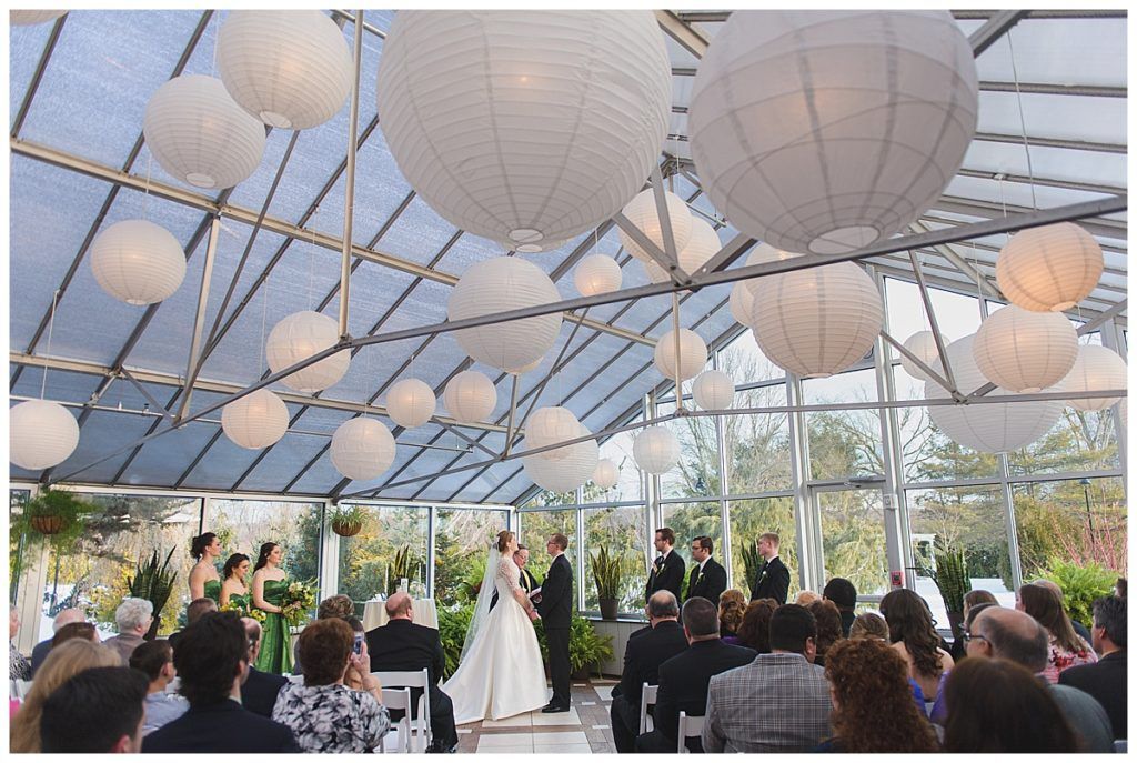 Wedding ceremony in a greenhouse with white paper lanterns. Bride, groom, and guests are present.