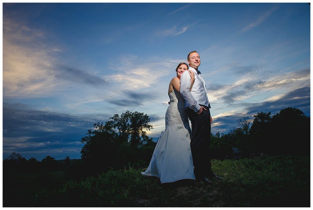 Bride and groom embracing outdoors at dusk, leaning backs, happy expressions, blue sky.