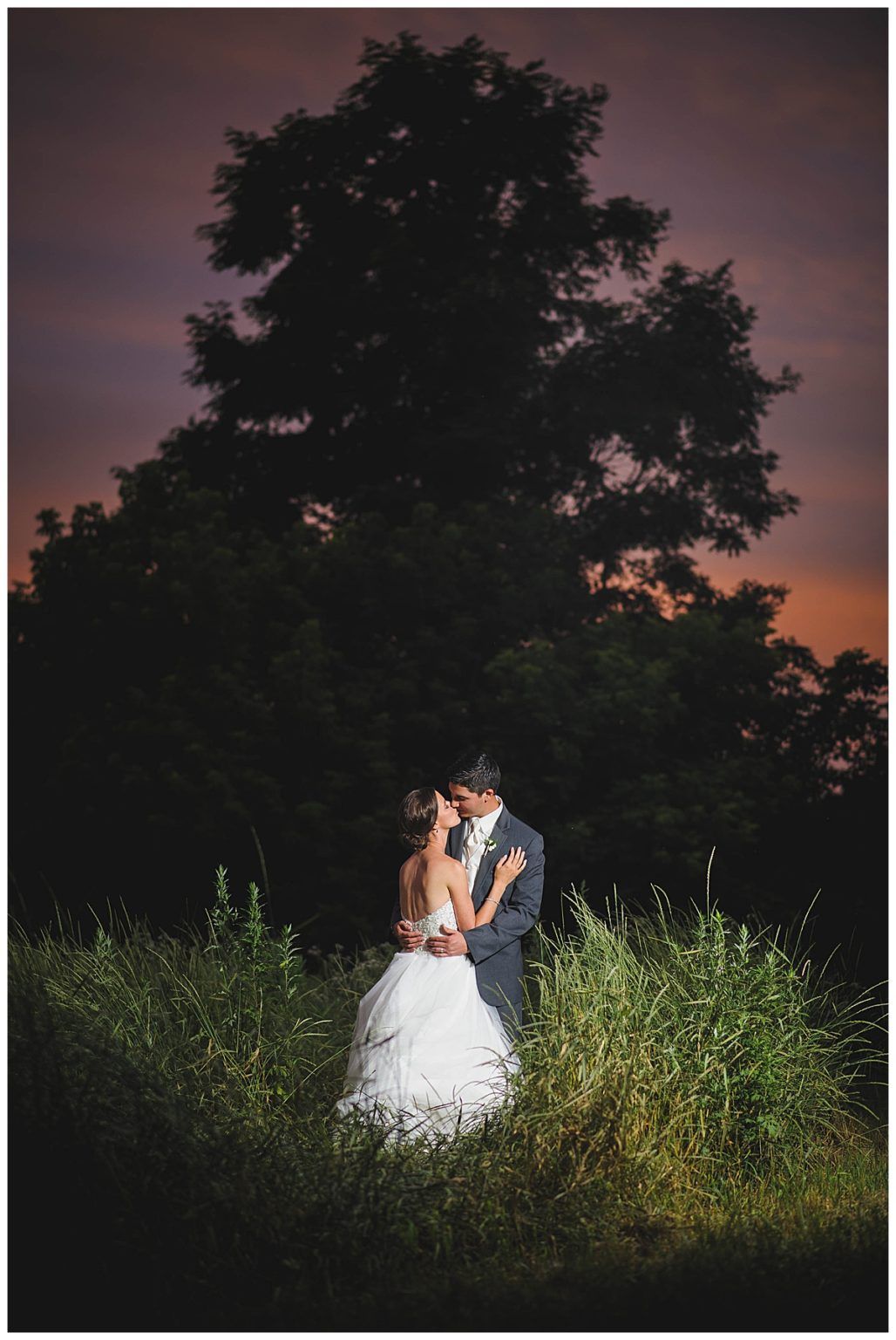 Couple embracing in wedding attire beneath a large tree, sunset sky in background.