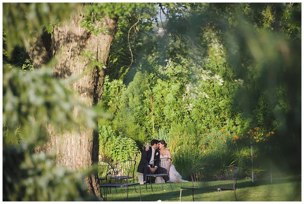 A couple sits by a fire pit surrounded by green trees and foliage in a sunlit outdoor setting.