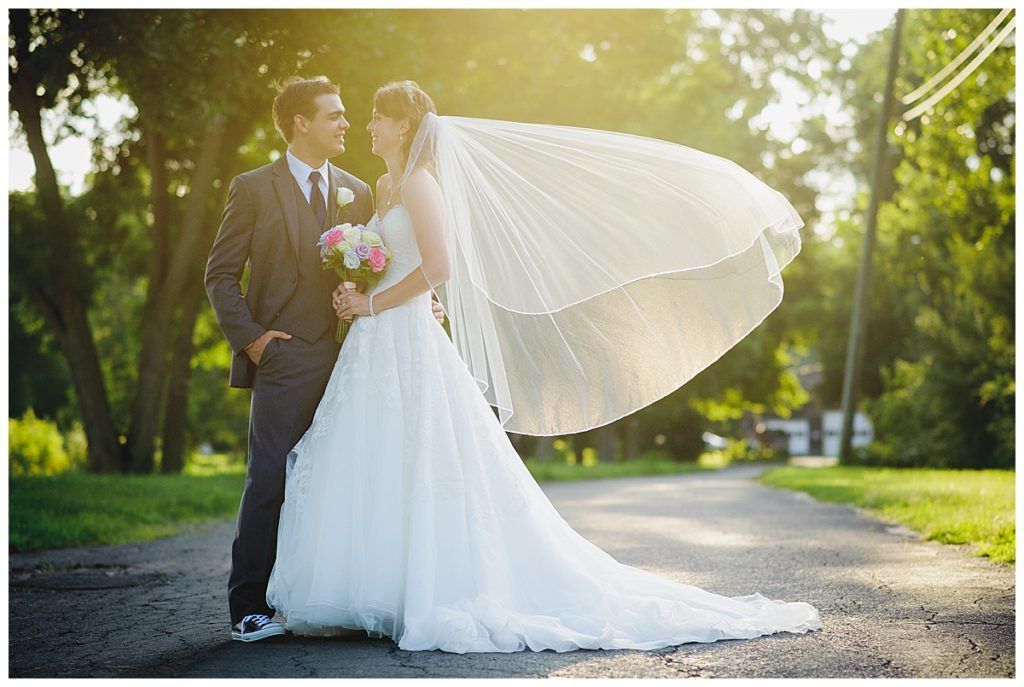 Bride and groom facing each other on a paved road, the bride's veil blowing in the wind. Sunlight filters through the trees.