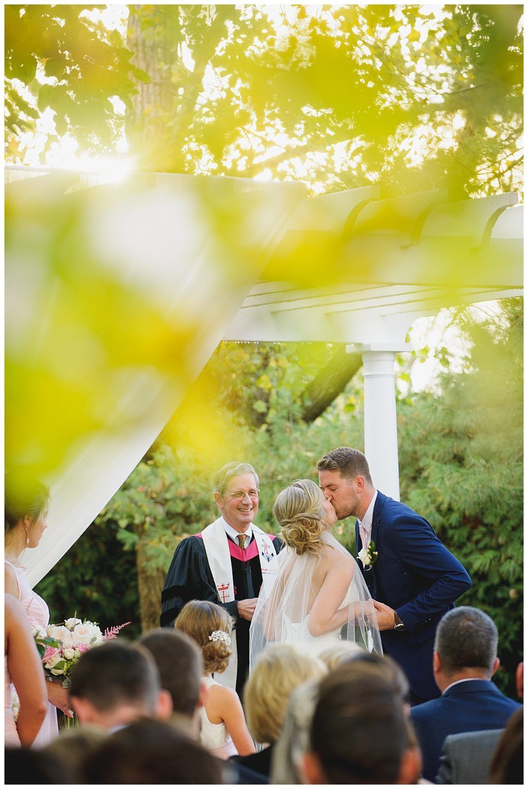 Wedding ceremony: couple kissing under a white arbor, officiant smiling. Guests seated. Sunlight filters through trees.