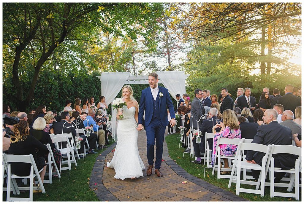 Newly married couple walks down aisle, surrounded by guests at outdoor ceremony.