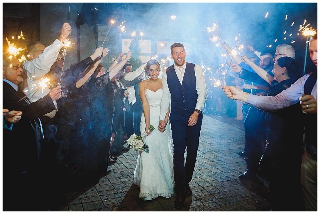 Newlyweds walk through sparkler-lined path, smiling. Wedding guests celebrate. Nighttime setting with stone floor.