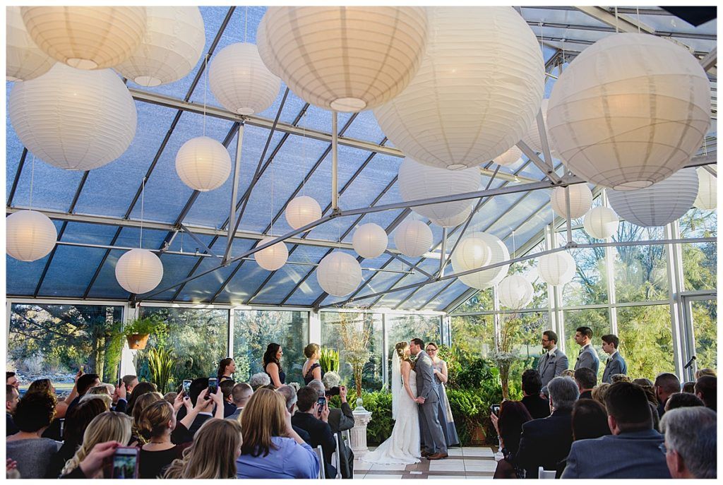 Wedding ceremony in a greenhouse with white paper lanterns. Bride and groom kissing on a small stage. Guests watch.