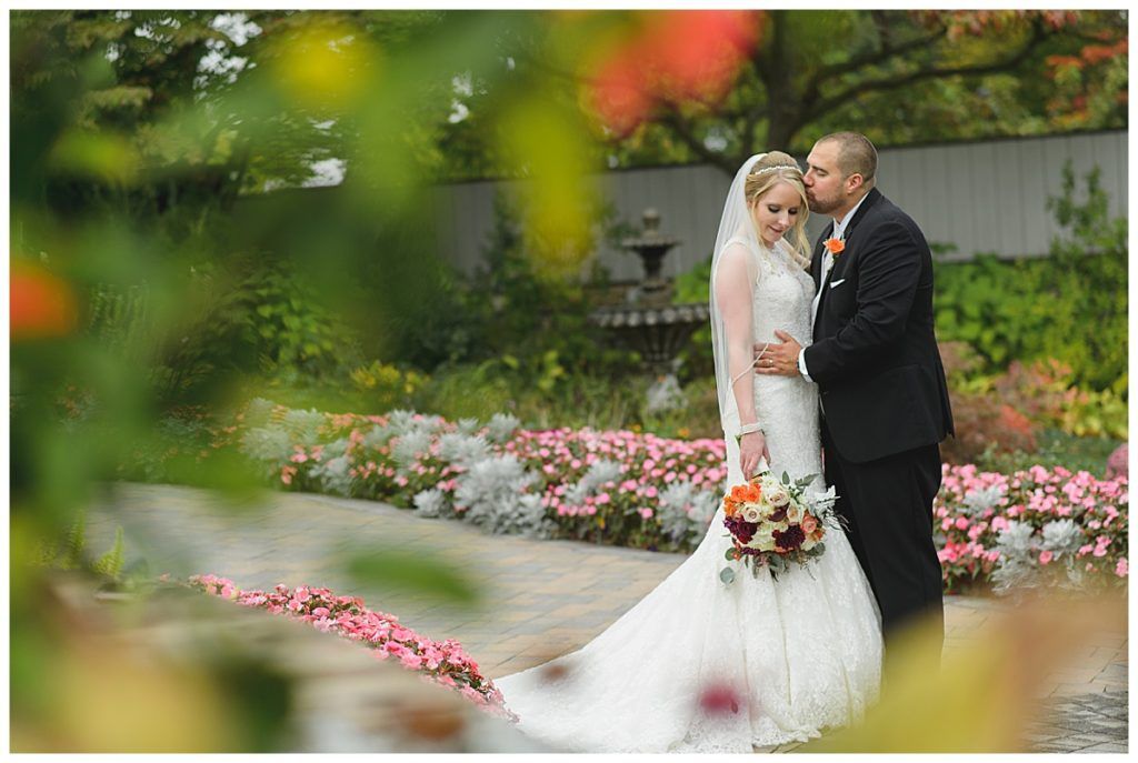 Bride and groom embrace in a garden. The groom kisses the bride's head, while holding her. Colorful flowers are in the background.