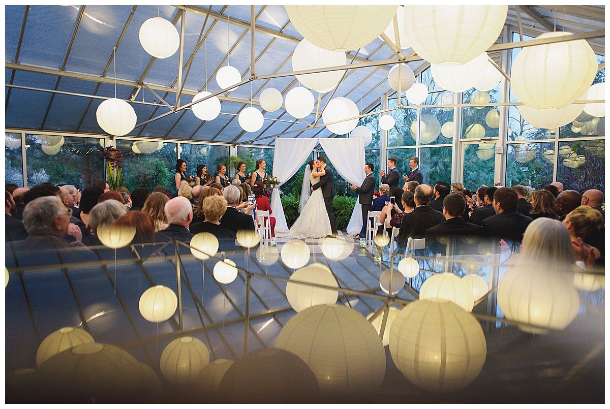 Wedding ceremony in a greenhouse with white paper lanterns. Bride and groom stand at the altar, guests seated.
