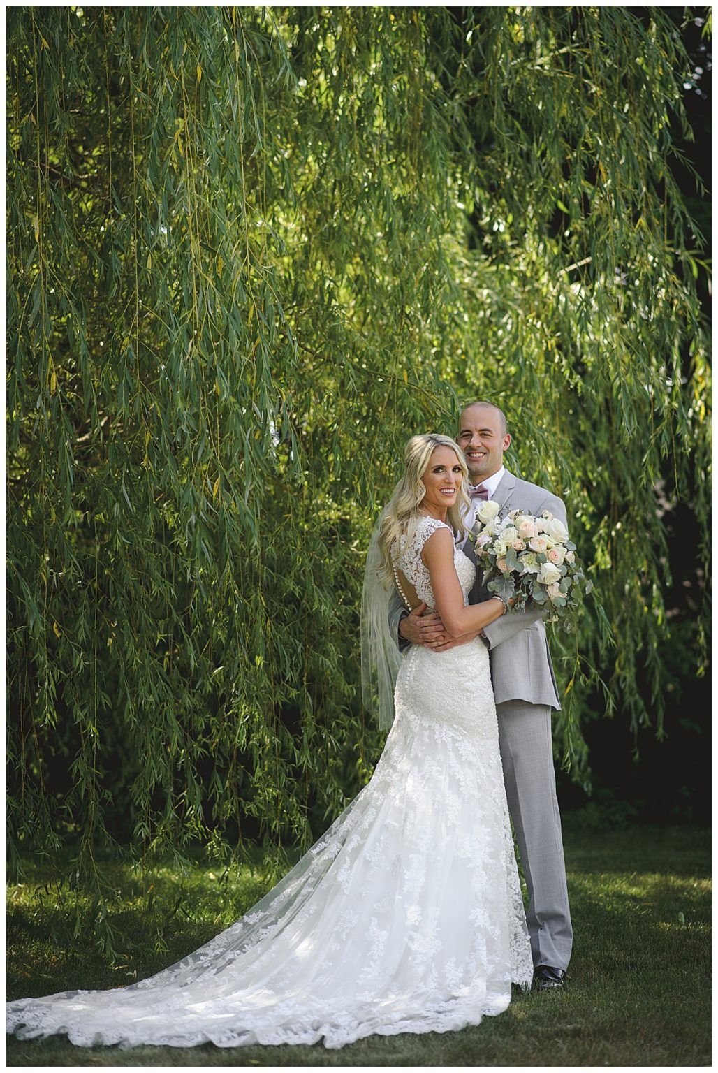 Bride and groom pose embracing, under a weeping willow. Bride in lace gown, holding bouquet. Groom in gray suit. Outdoors.