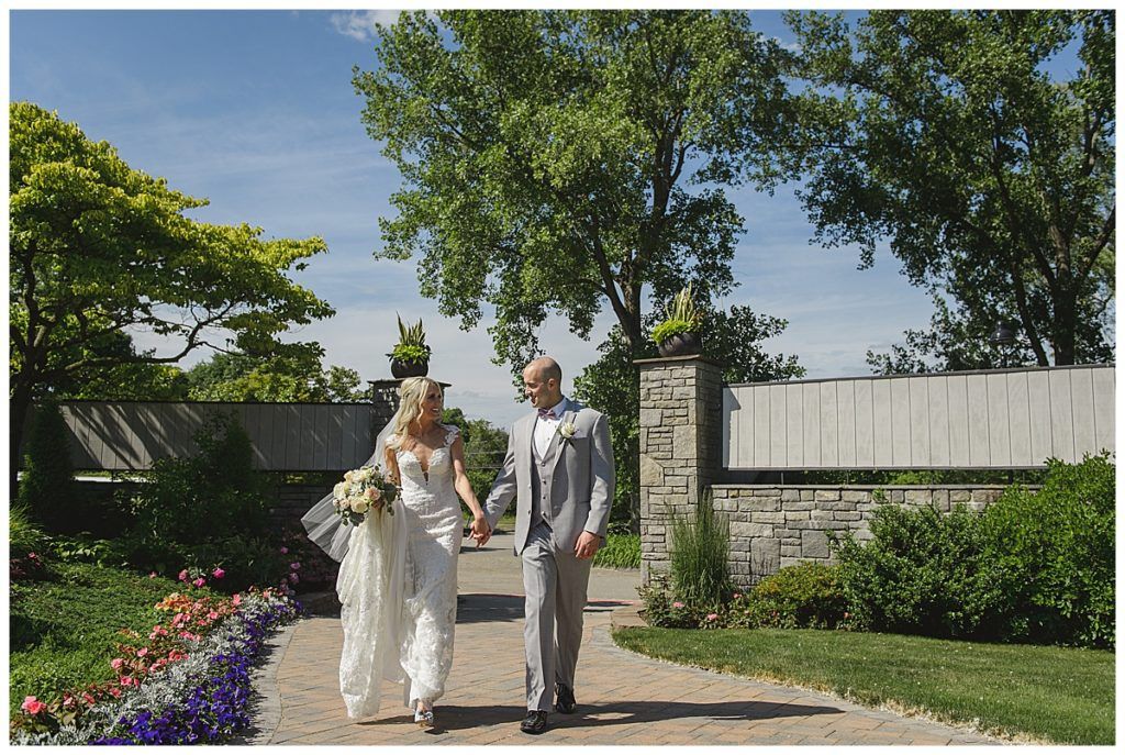 Bride and groom walking hand-in-hand on a sunny path, flowers and greenery surrounding.
