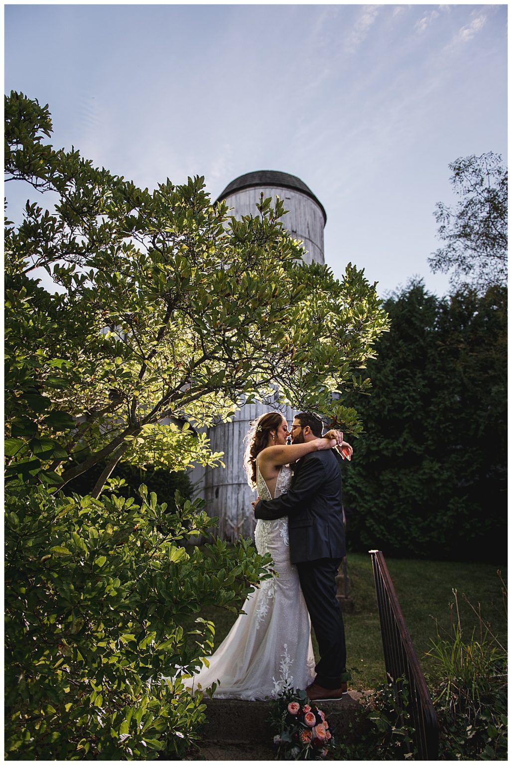 Couple kissing, silhouetted in front of a silo, beneath a tree, on their wedding day.
