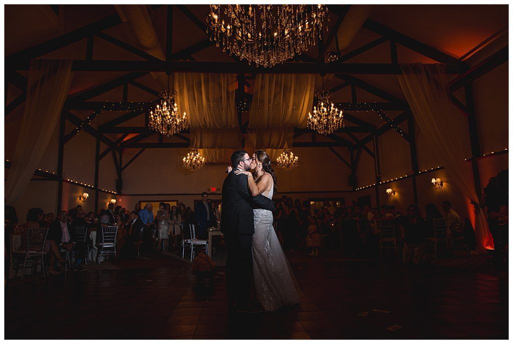Bride and groom share a kiss on the dance floor in a warmly lit barn, chandeliers overhead.