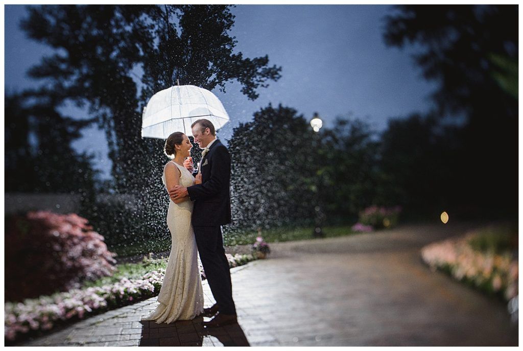 Bride and groom under an umbrella in the rain, on a brick path, with lit flowers and trees.
