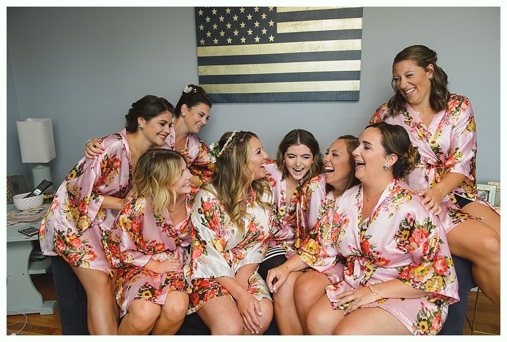 Bridesmaids in pink floral robes laugh together on a couch, American flag in background.